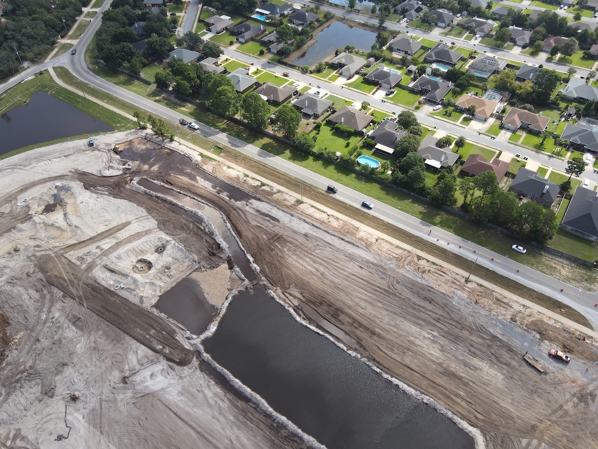 Aerial shot of a Green Energy Contracting job site near Panama City, FL showing cleared and graded land with preparations being made for underground utilities. Adjacent to the job site is a road with vehicles and behind that a few rows of houses